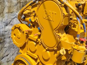 Close-up of a large yellow Caterpillar diesel engine component with blue fittings and hoses attached to a rock wall backdrop.
